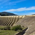 Vista ampla de uma grande barragem de concreto sob céu azul, representando infraestrutura hídrica.