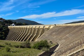Vista ampla de uma grande barragem de concreto sob céu azul, representando infraestrutura hídrica.